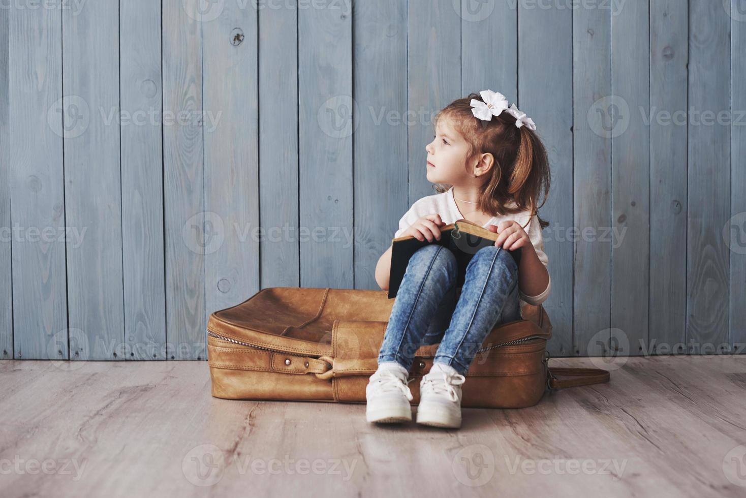 Ready to big travel. Happy little girl reading interesting book carrying a big briefcase and smiling. Travel, freedom and imagination concept photo