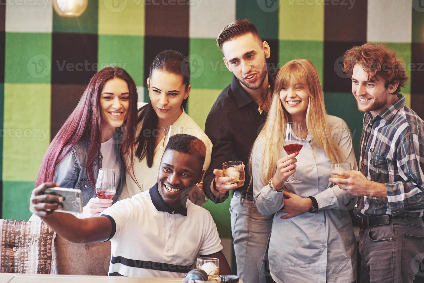 amigos divirtiéndose en el restaurante dos niños y cuatro niñas bebiendo haciendo selfie, haciendo el signo de la paz y riendo. en primer plano mujer sosteniendo teléfono inteligente. todos usan ropa casual foto