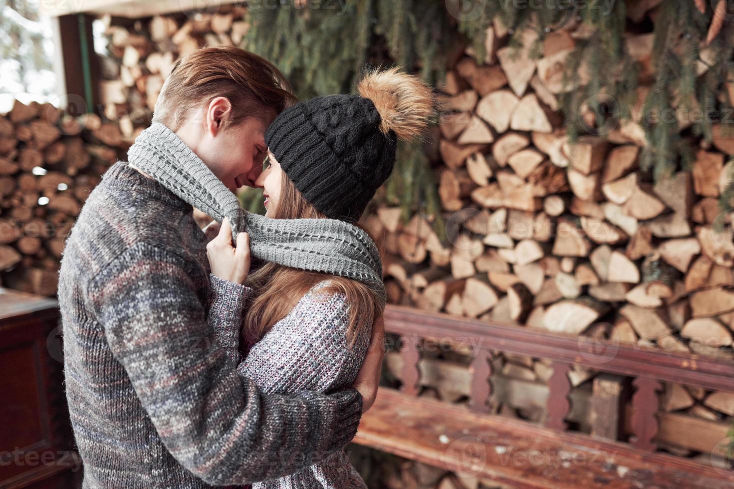 Concepto de invierno, vacaciones, pareja, Navidad y personas - hombre y mujer sonrientes con sombreros y bufanda abrazándose sobre casa de campo de madera y fondo de nieve foto