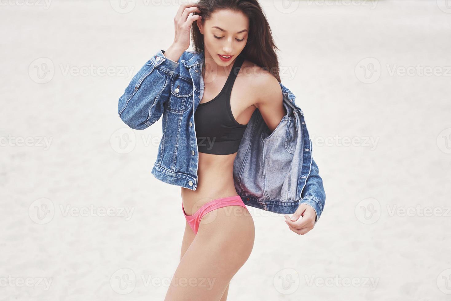 Beautiful slim luxury girl in bikini and jeans jaket on the sand beach on a tropical island. Sexy tanned body and perfect figure photo