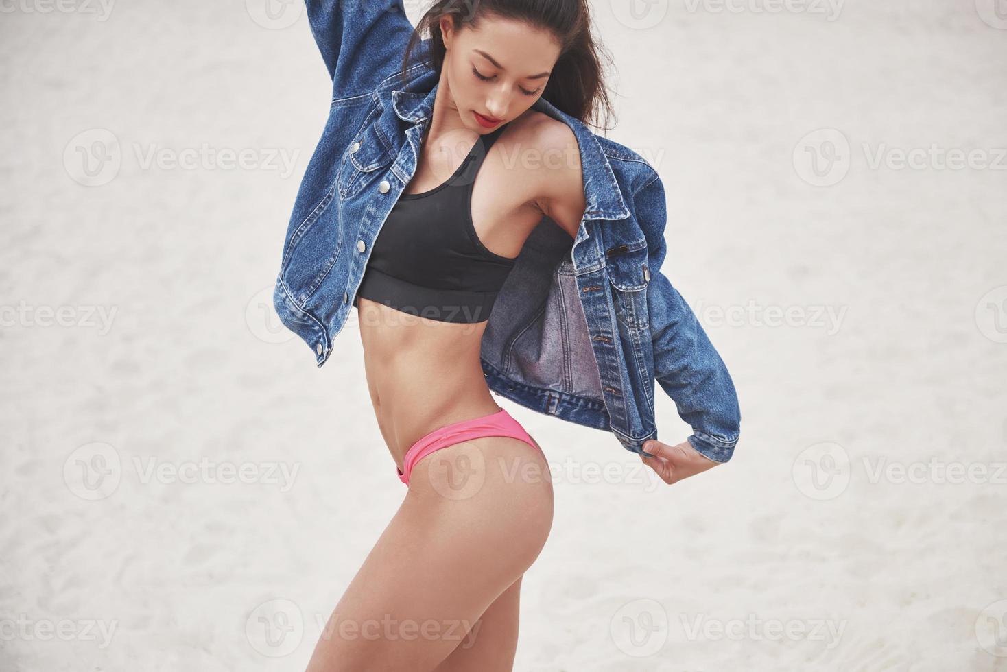 hermosa chica de lujo delgada en bikini y jeans jaket en la playa de arena en una isla tropical. sexy cuerpo bronceado y figura perfecta foto