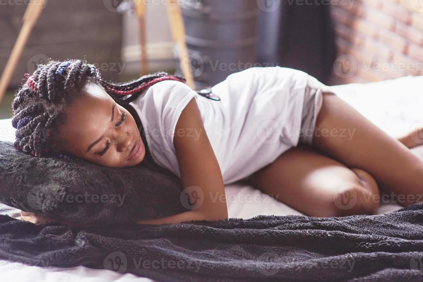 A beautiful African woman lies and sleeps in bed, in a white bedroom. Good morning, vacation at home. The girl is wearing a T-shirt, pigtails on her head photo