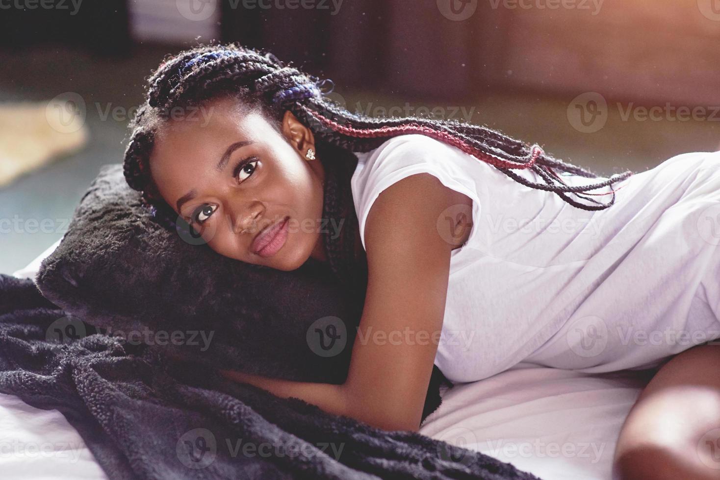 A beautiful African woman lies and sleeps in bed, in a white bedroom. Good morning, vacation at home. The girl is wearing a T-shirt, pigtails on her head photo