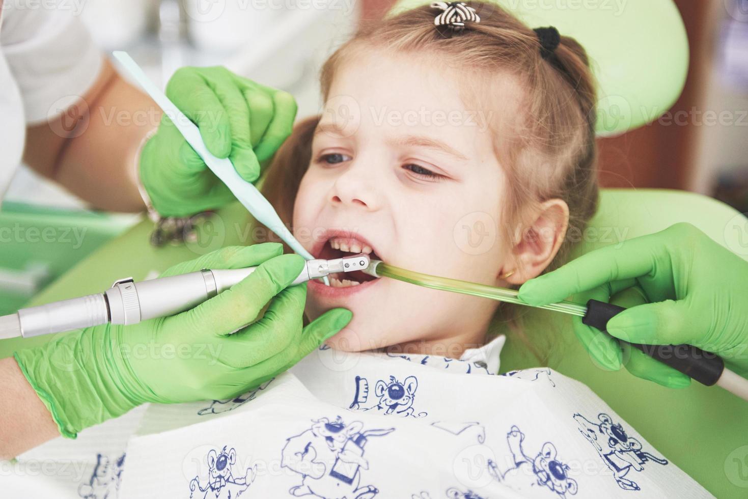 Manos de dentista pediátrico irreconocible y asistente que hace el procedimiento de examen para una niña linda sonriente sentada en una silla en el hospital foto