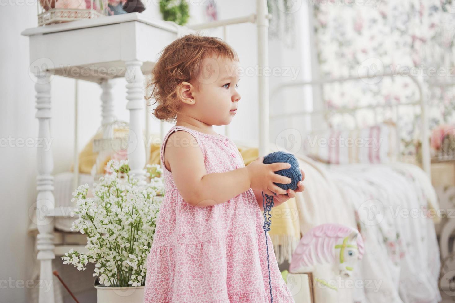 Childhood concept. Baby girl in cute dress play with colored thread. White vintage childroom photo