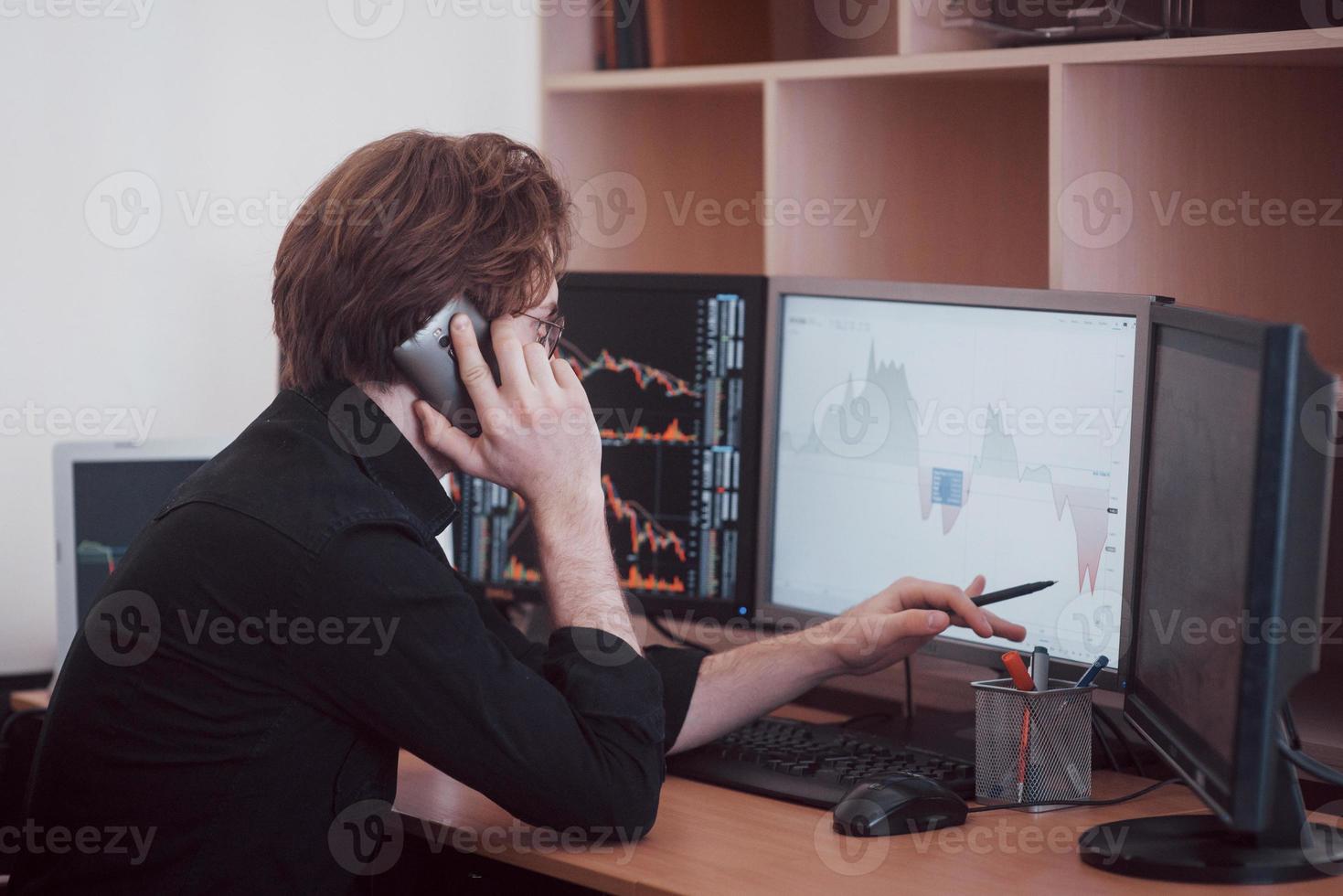 Over the shoulder view of and stock broker trading online while accepting orders by phone. Multiple computer screens ful of charts and data analyses in background photo