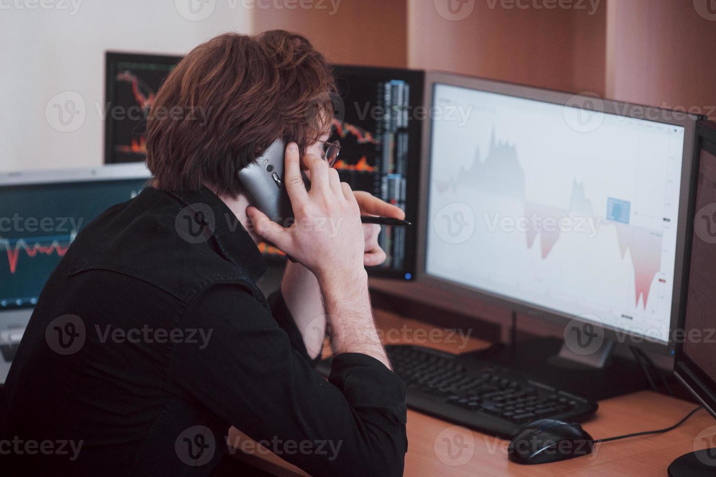 Over the shoulder view of and stock broker trading online while accepting orders by phone. Multiple computer screens ful of charts and data analyses in background photo