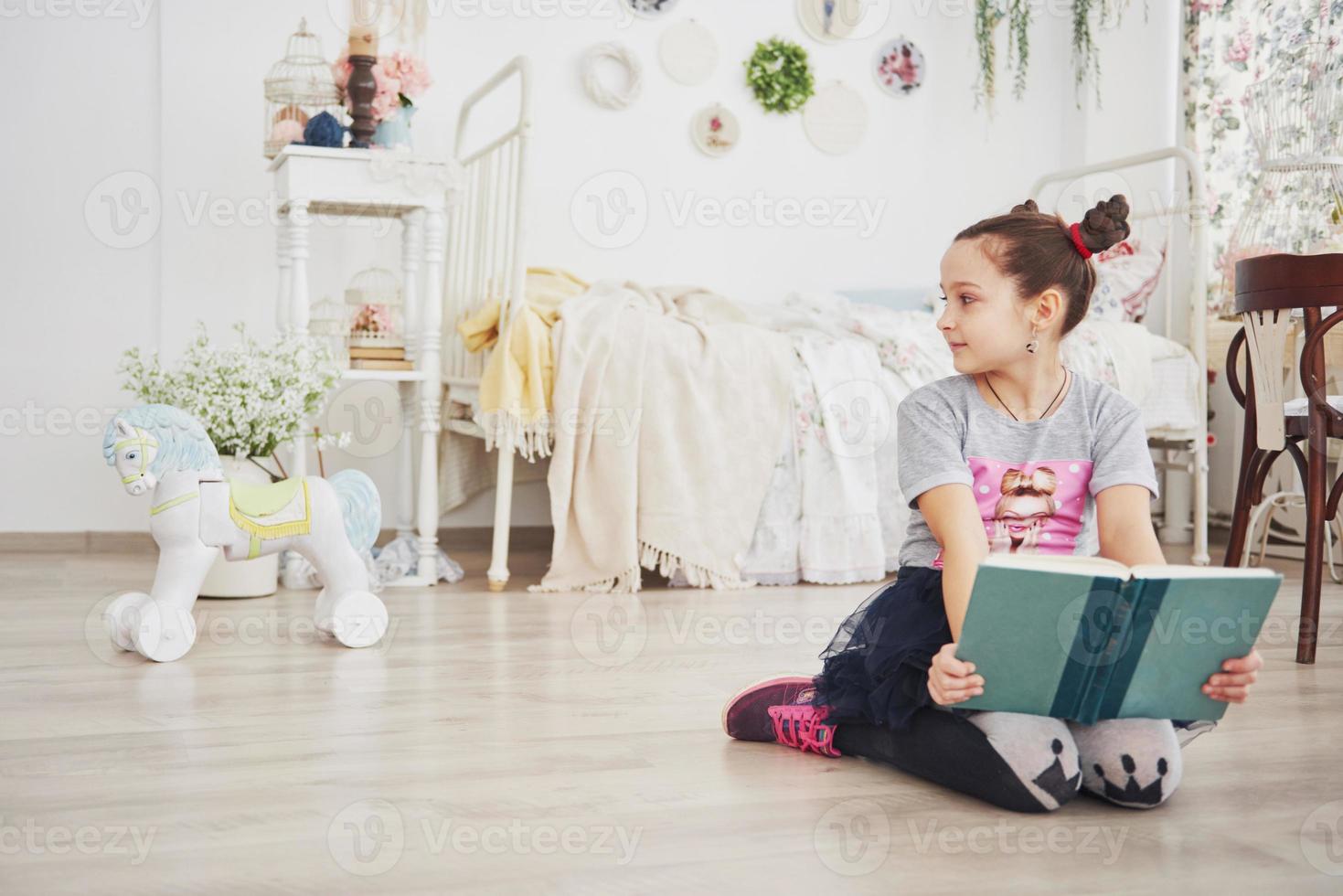 niña linda niña leyendo un libro en el dormitorio. niño con corona sentado en la cama cerca de la ventana foto