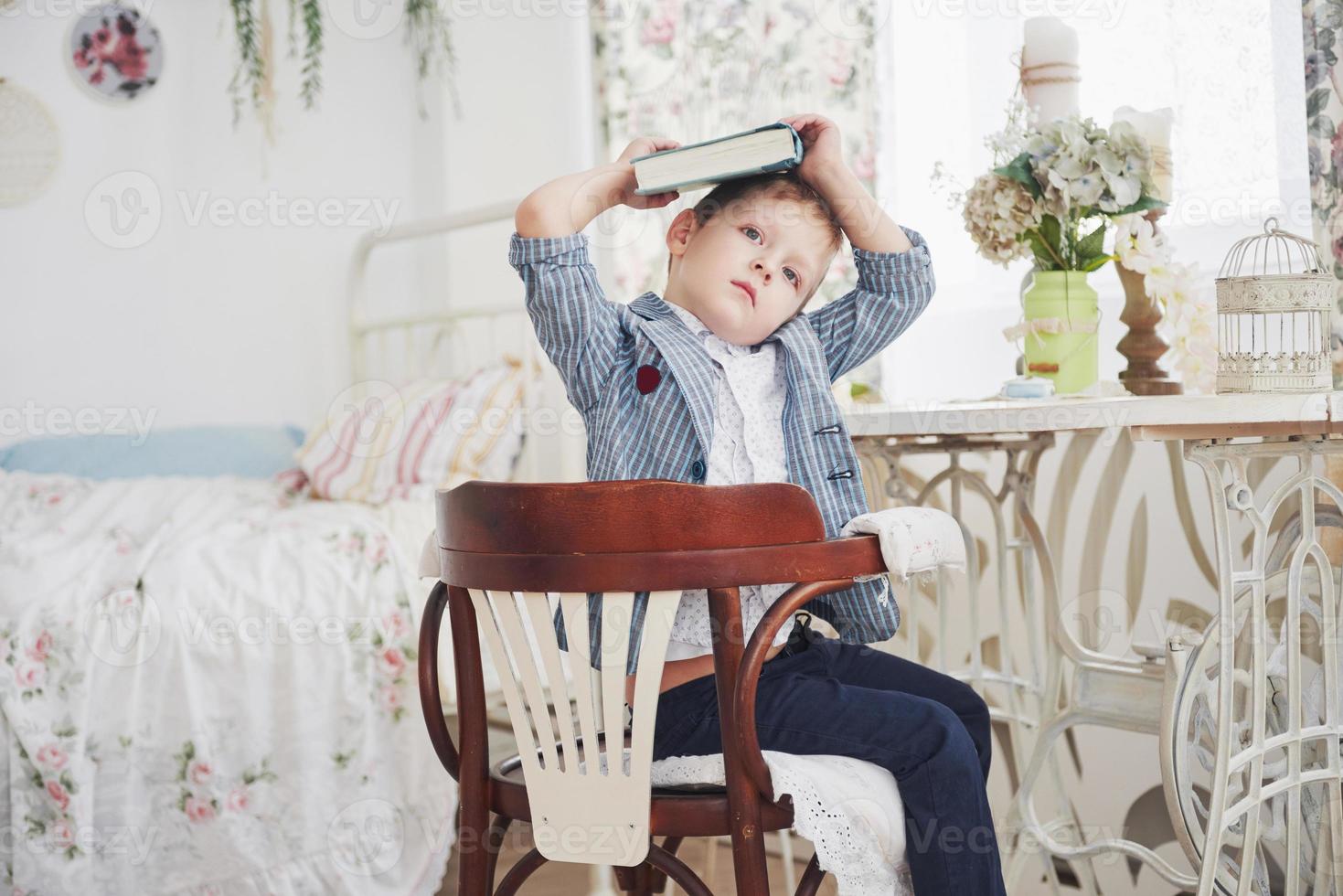 Photo of diligent schoolboy with book on his head doing homework. The schoolboy is tired of doing homework