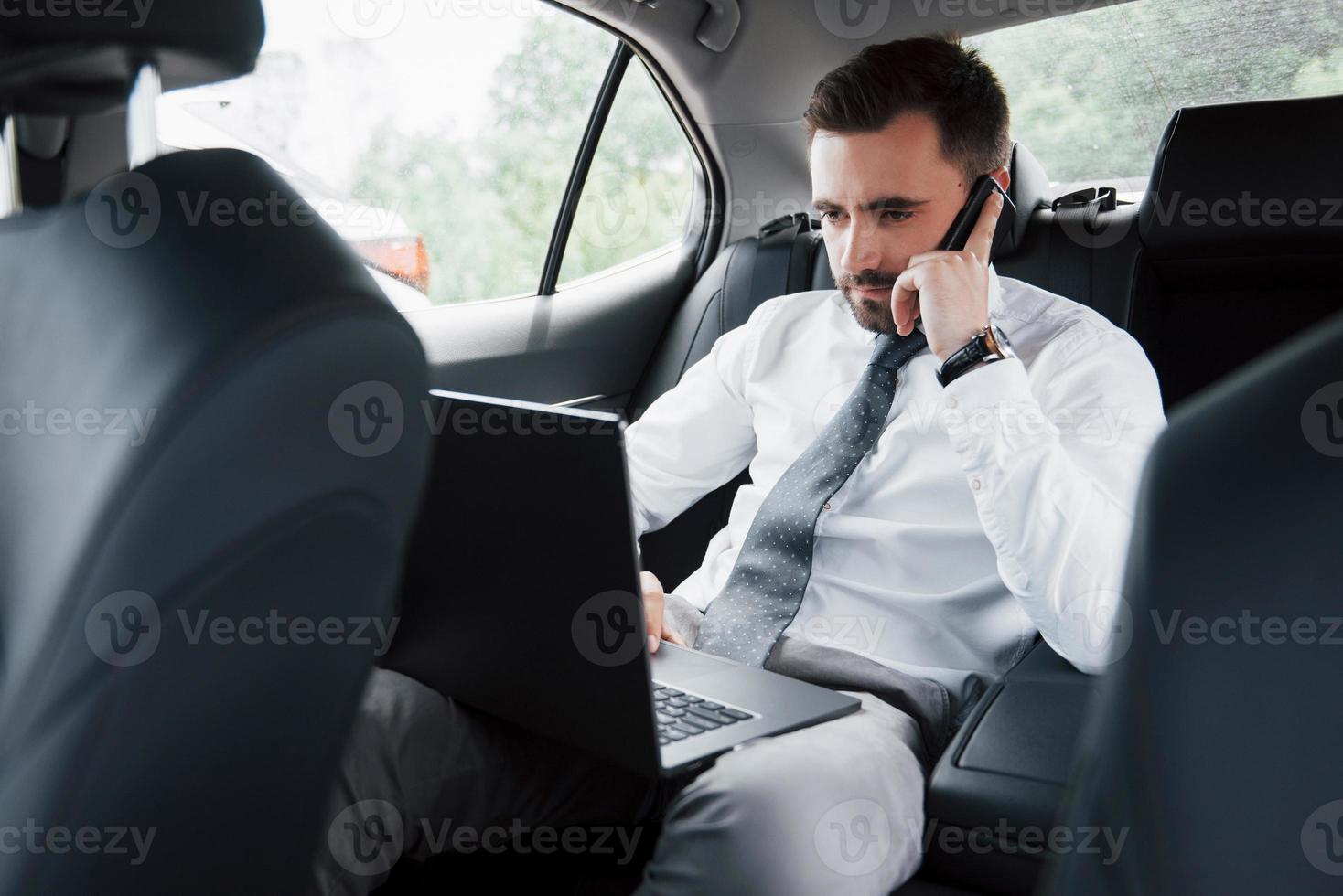 A young businessman working on laptop and talking on the phone while sitting in the car's back. Works in motion, appreciates its time photo