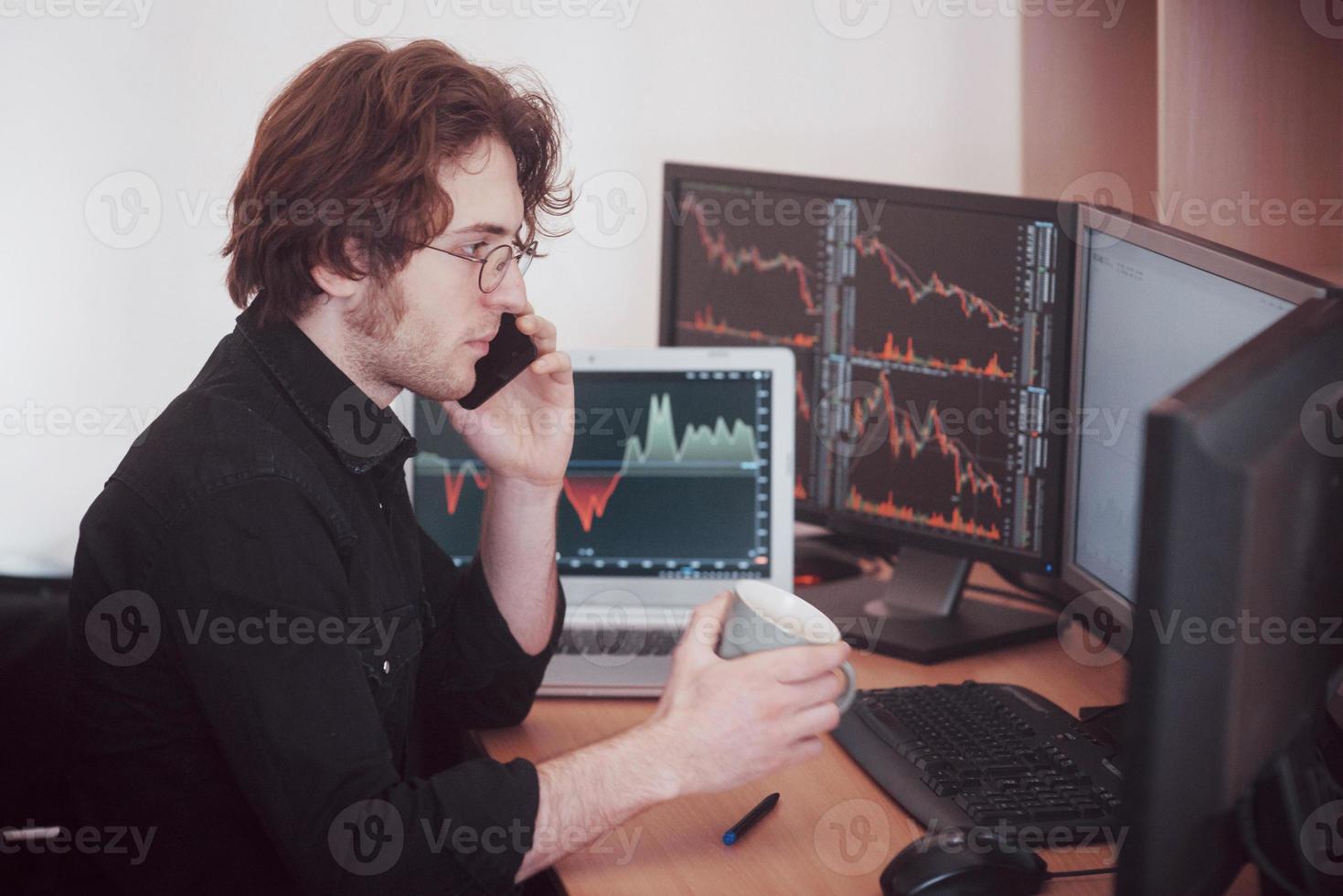 Over the shoulder view of and stock broker trading online while accepting orders by phone. Multiple computer screens ful of charts and data analyses in background photo