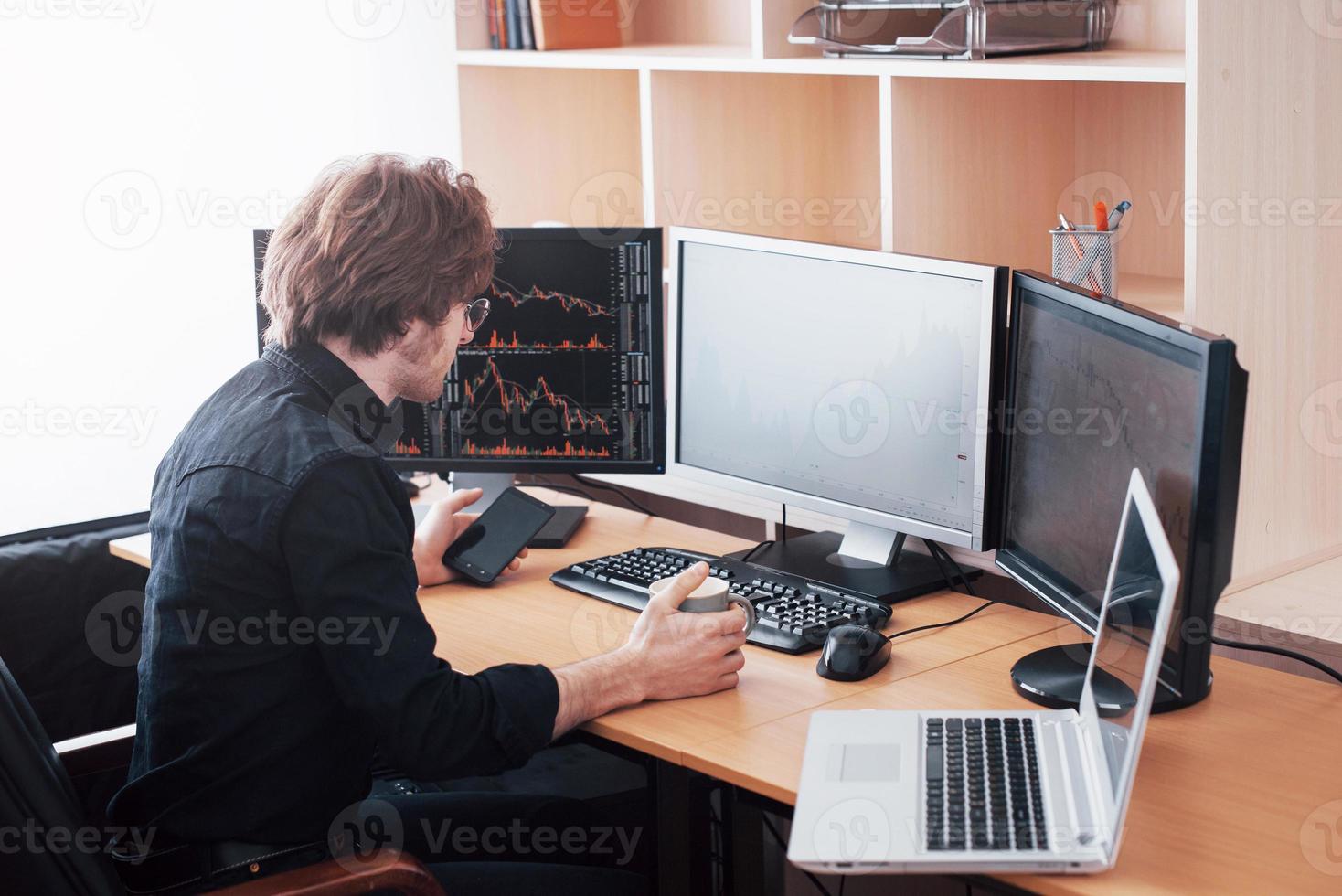 Over the shoulder view of and stock broker trading online while accepting orders by phone. Multiple computer screens ful of charts and data analyses in background photo