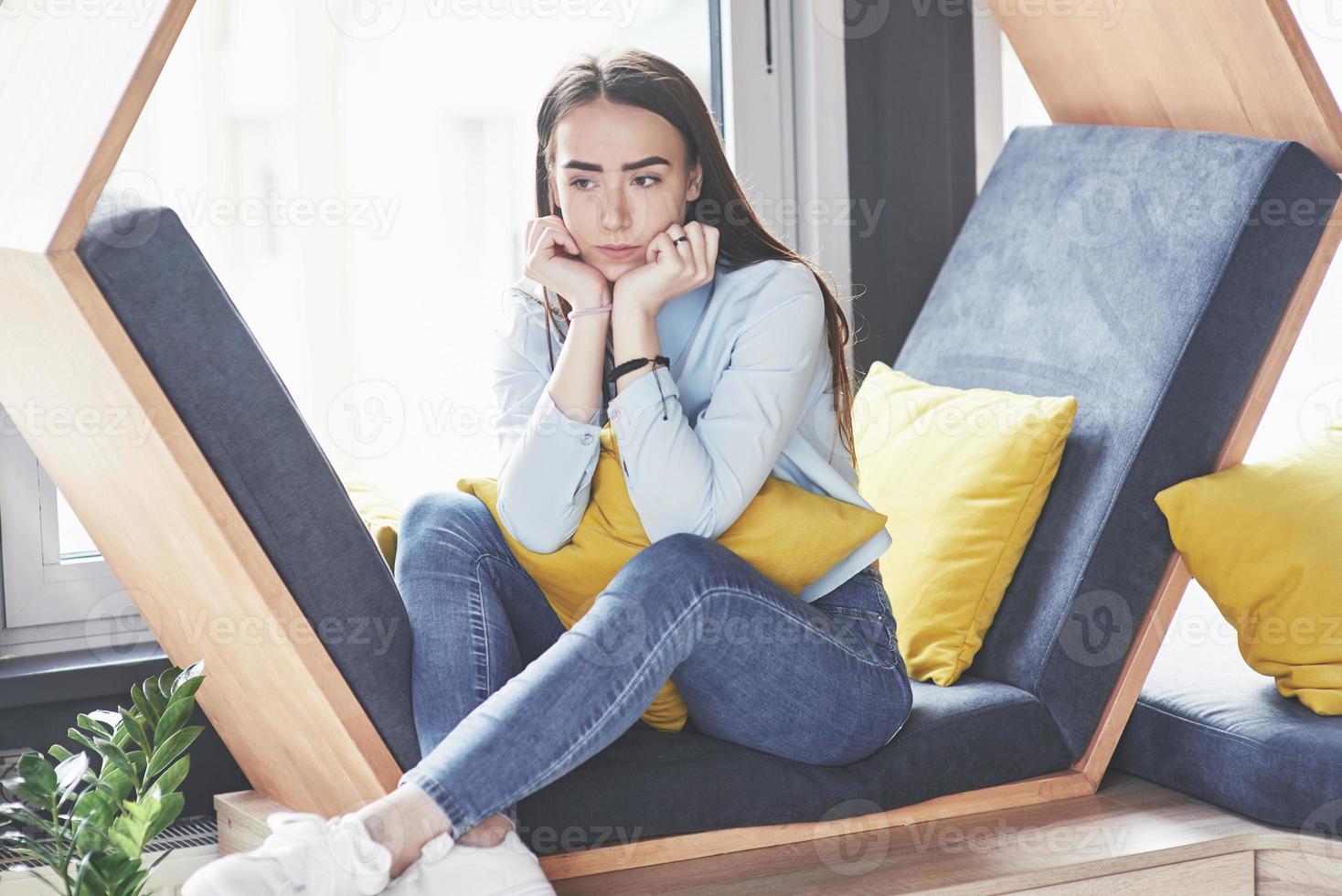 Shy cute girl spends her time in the recreation area. She sits and has fun in the sofa chair in the form of a hexagon with pillows near the window photo