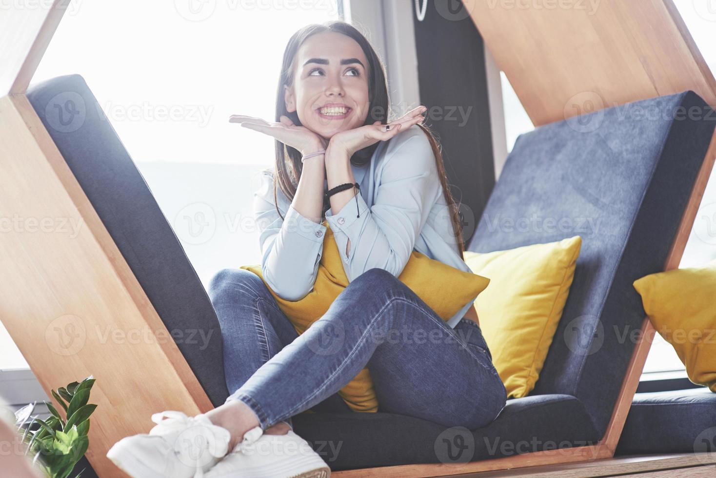 Shy cute girl spends her time in the recreation area. She sits and has fun in the sofa chair in the form of a hexagon with pillows near the window photo