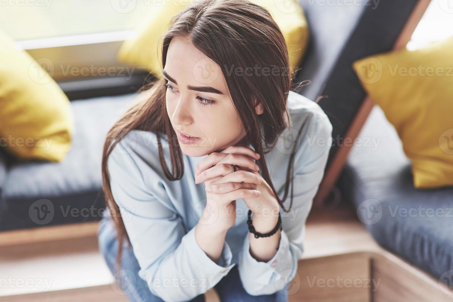 Beautiful young girl spends her time in the recreation area. She sits and has fun in the sofa chair in the form of a hexagon with pillows near the window photo