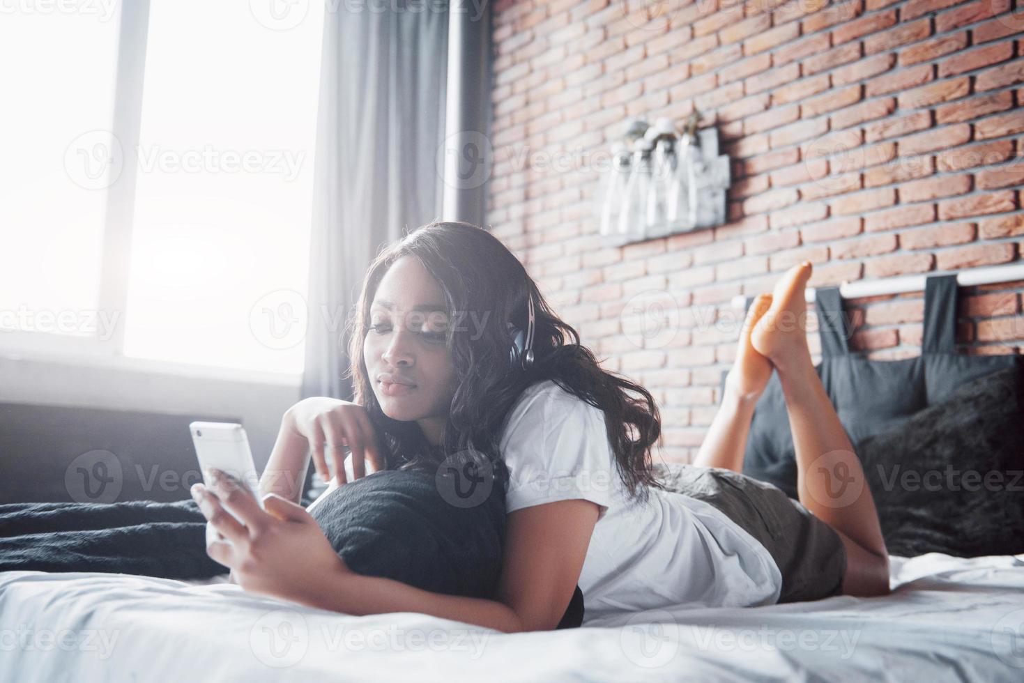 Hermosa niña africana en ropa de dormir sonriendo escuchando música en auriculares acostado en la cama foto