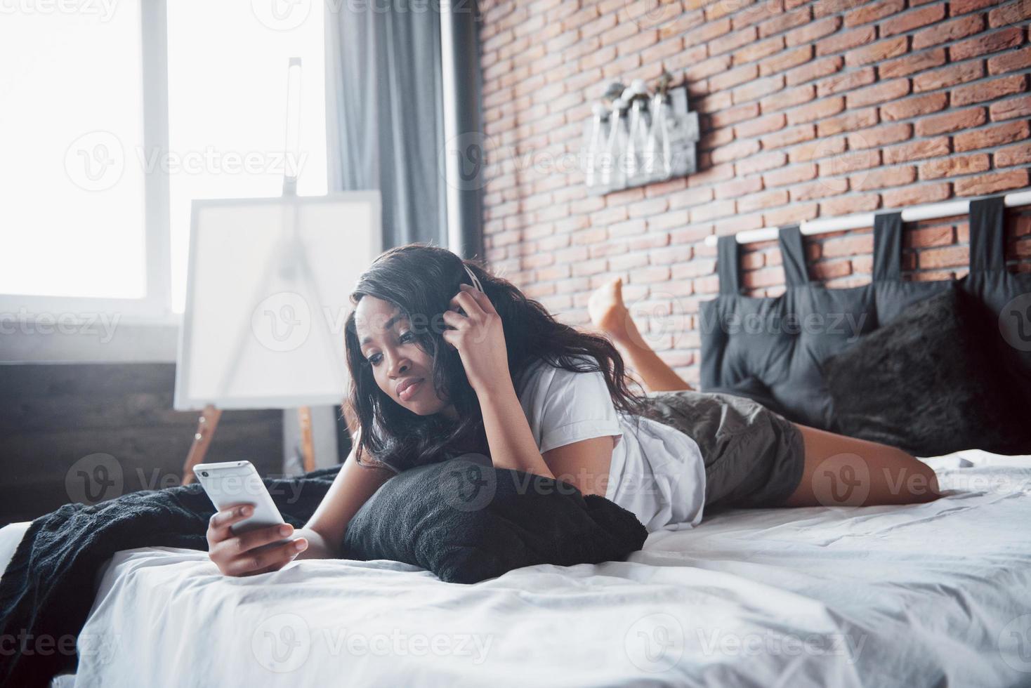 Hermosa niña africana en ropa de dormir sonriendo escuchando música en auriculares acostado en la cama foto