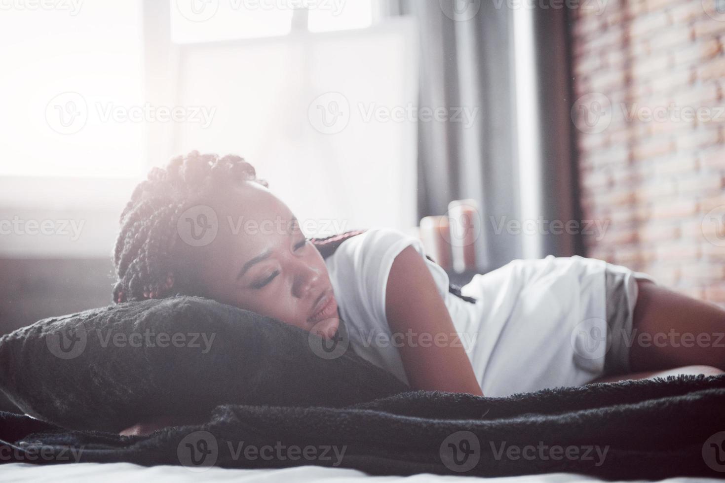 una hermosa mujer africana yace y duerme en la cama, en un dormitorio blanco. buenos dias vacaciones en casa. la niña lleva una camiseta, coletas en la cabeza foto