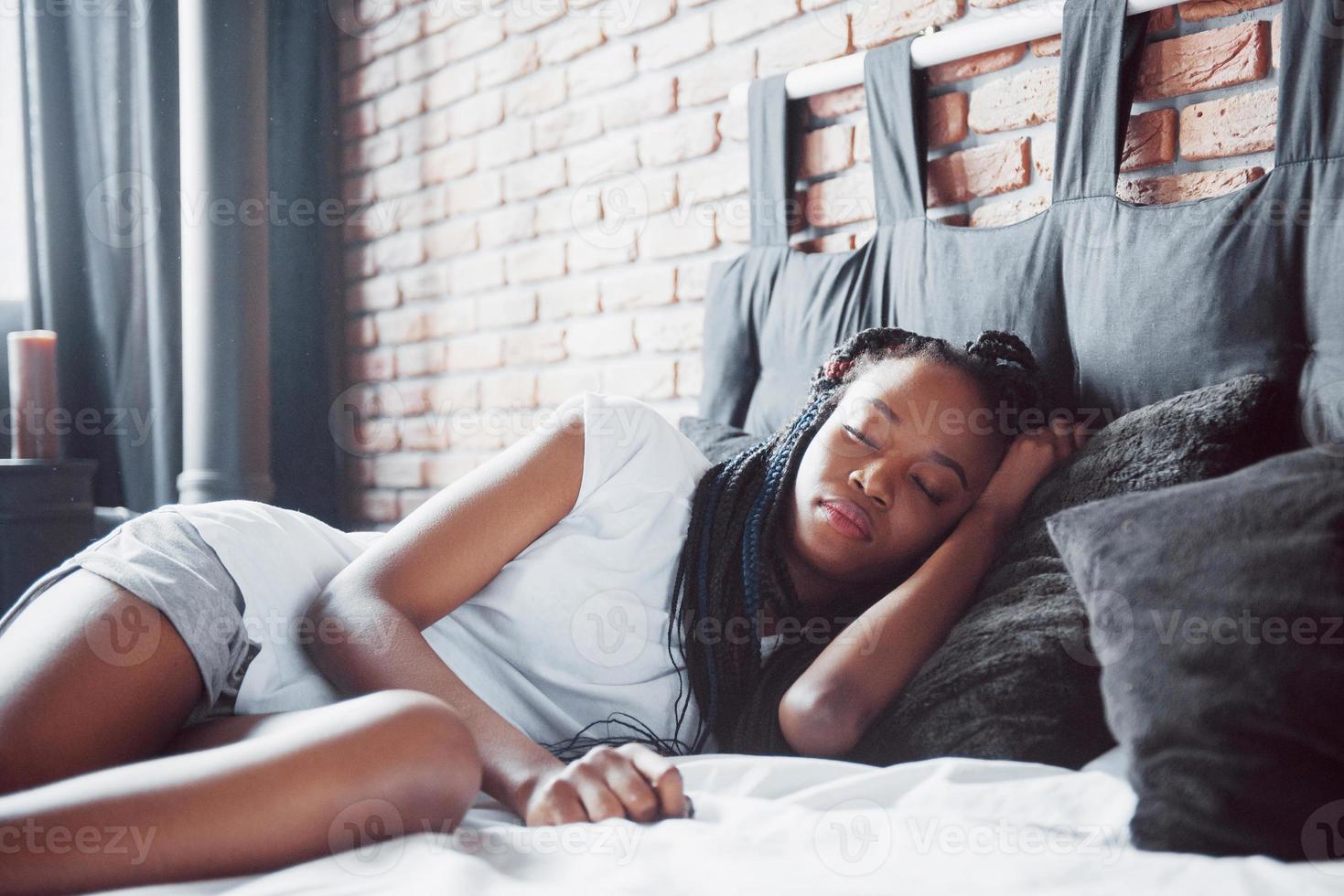 una hermosa mujer africana yace y duerme en la cama, en un dormitorio blanco. buenos dias vacaciones en casa. la niña lleva una camiseta, coletas en la cabeza foto