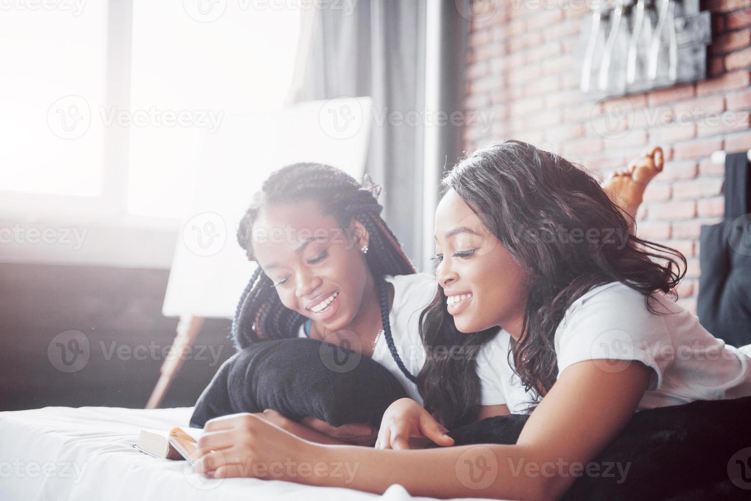 Two beautiful african girl in sleepwear smiling sitting on bed at home woke up in the morning on a sunny day. photo