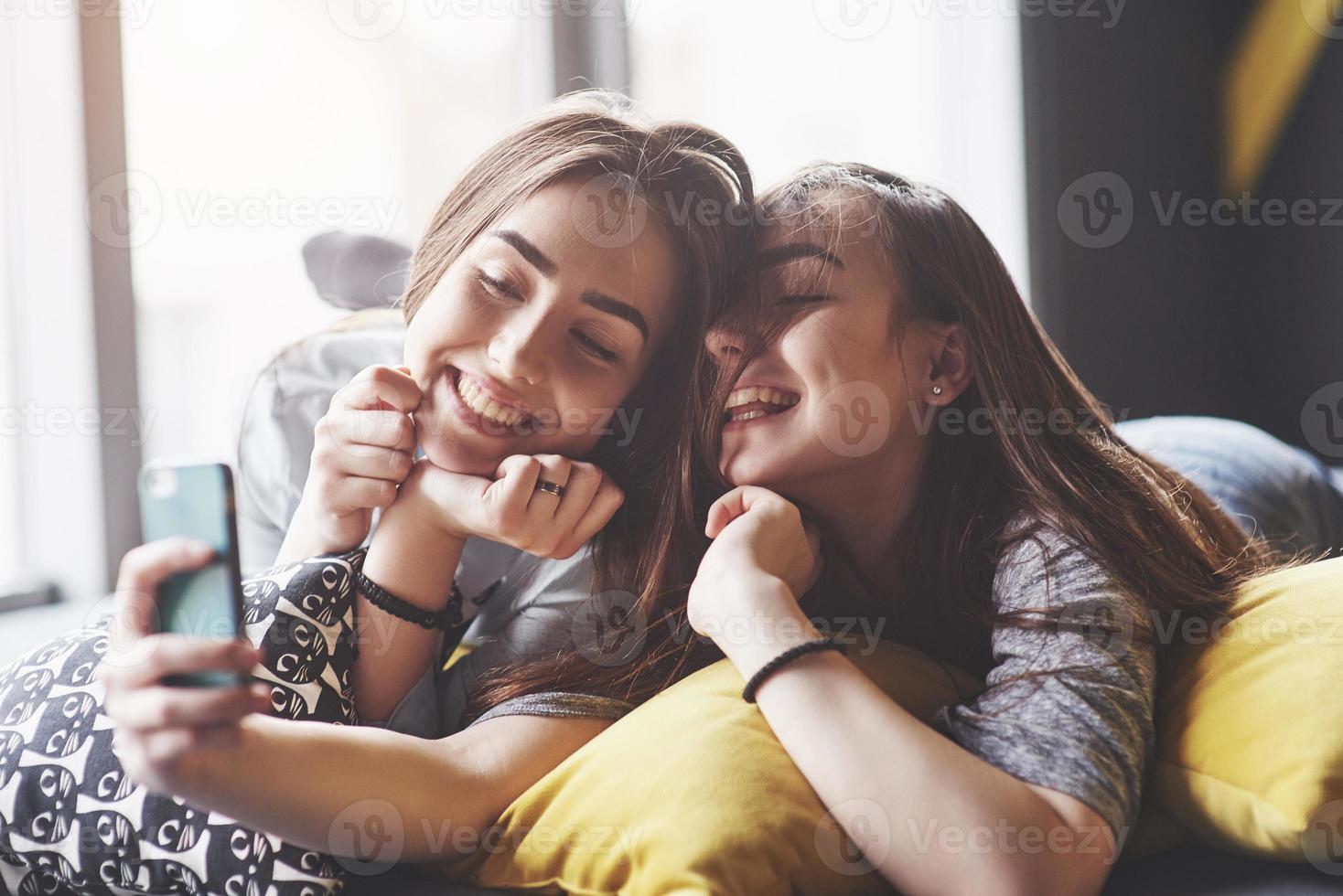 Two cute smiling twins sisters holding smartphone and making selfie. Girls lie on the couch posing and joy photo