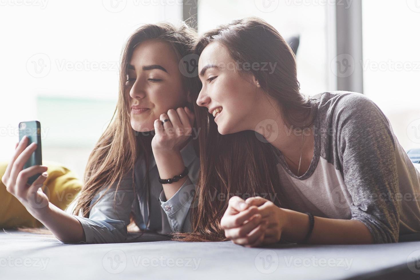 dos lindas hermanas gemelas sonrientes sosteniendo smartphone y haciendo selfie. las niñas se acuestan en el sofá posando y alegría foto