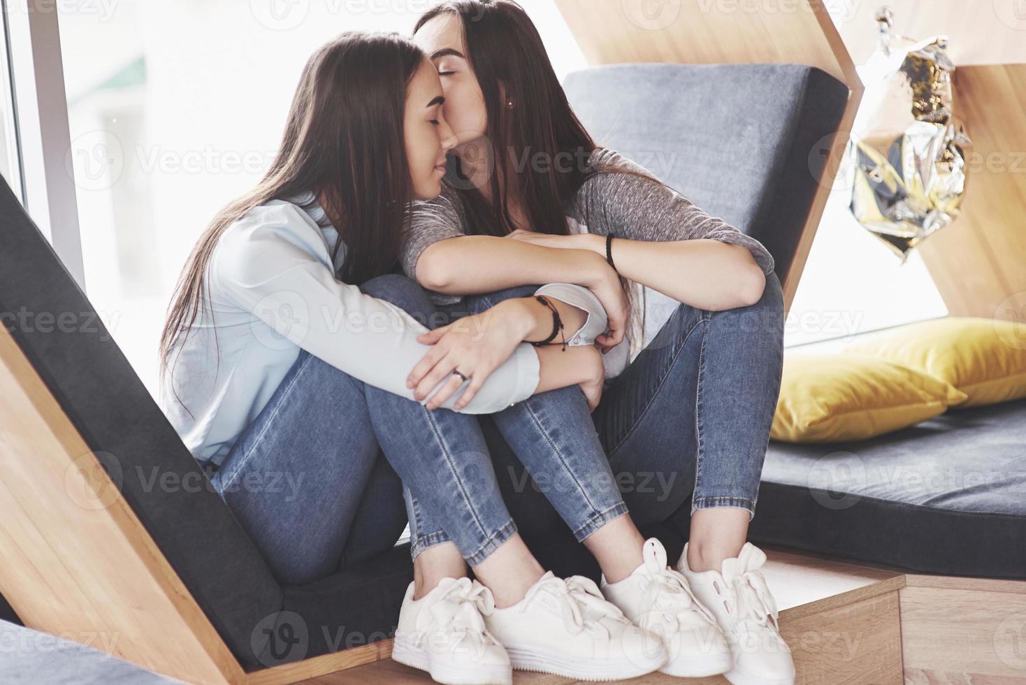 Two twin girls sit in a tight armchair for them in the recreation area. Sisters posing and having fun spend time. Always join friendship and youth concept photo