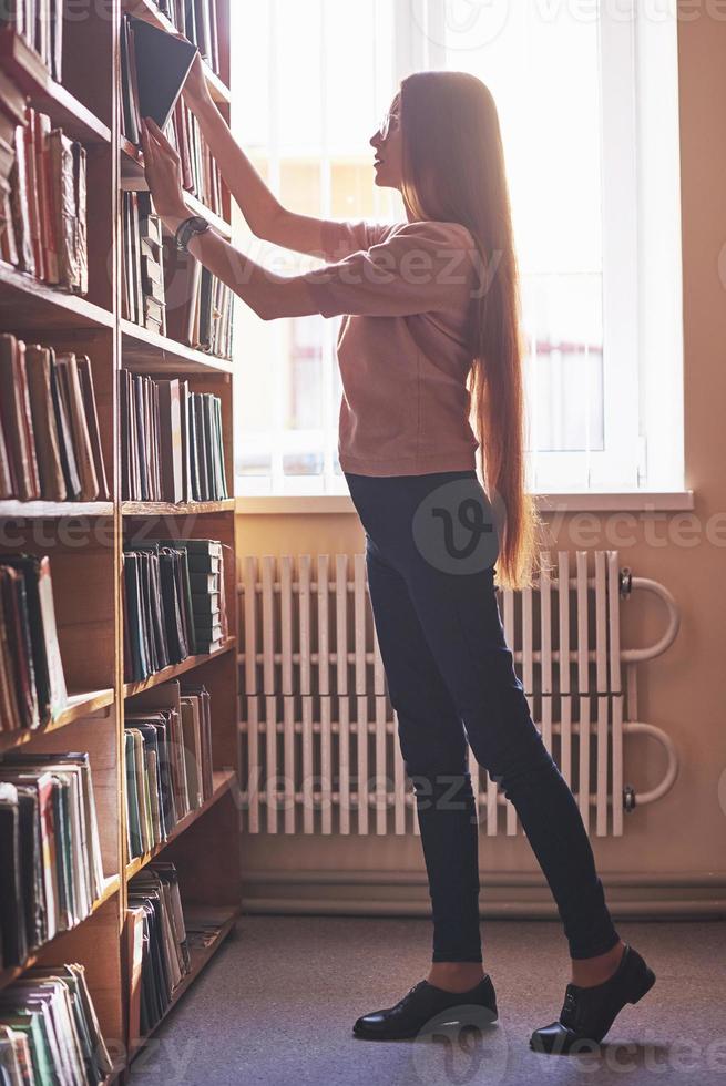 A young student girl is looking for the right book on the shelves of the old university library photo