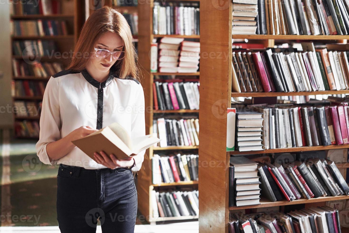 Joven y atractiva bibliotecaria estudiante leyendo un libro entre las estanterías de la biblioteca foto