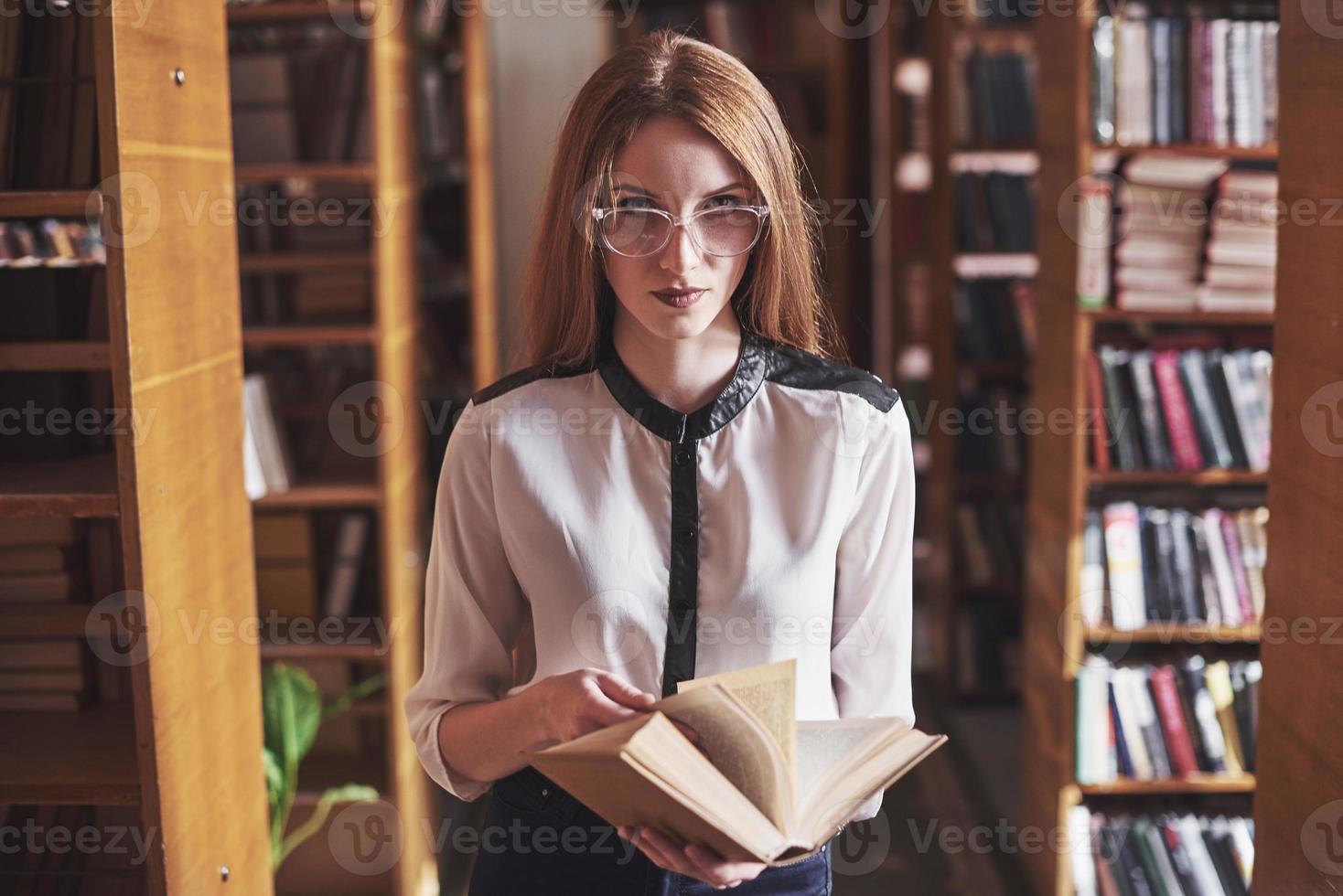 Joven y atractiva bibliotecaria estudiante leyendo un libro entre las estanterías de la biblioteca foto