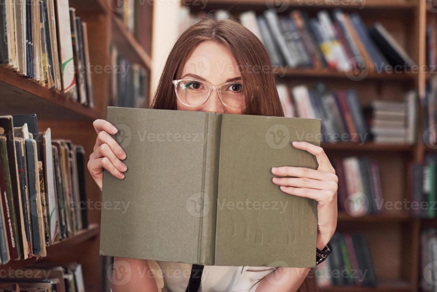 Young attractive student librarian reading a book between library bookshelves photo