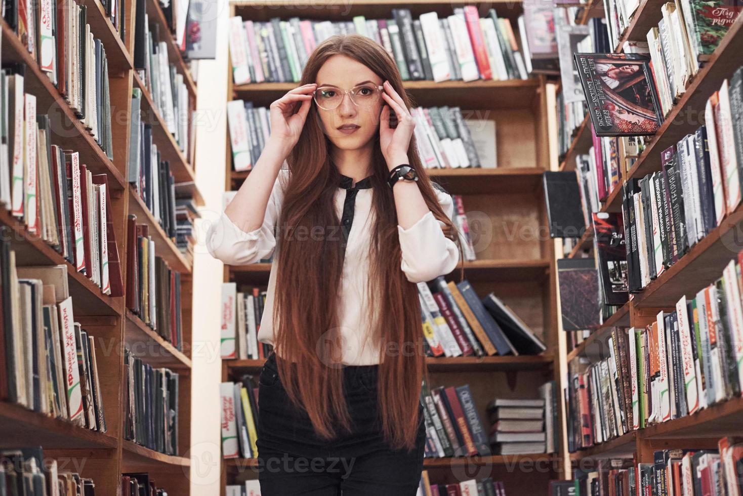 Joven y atractiva bibliotecaria estudiante leyendo un libro entre las estanterías de la biblioteca foto