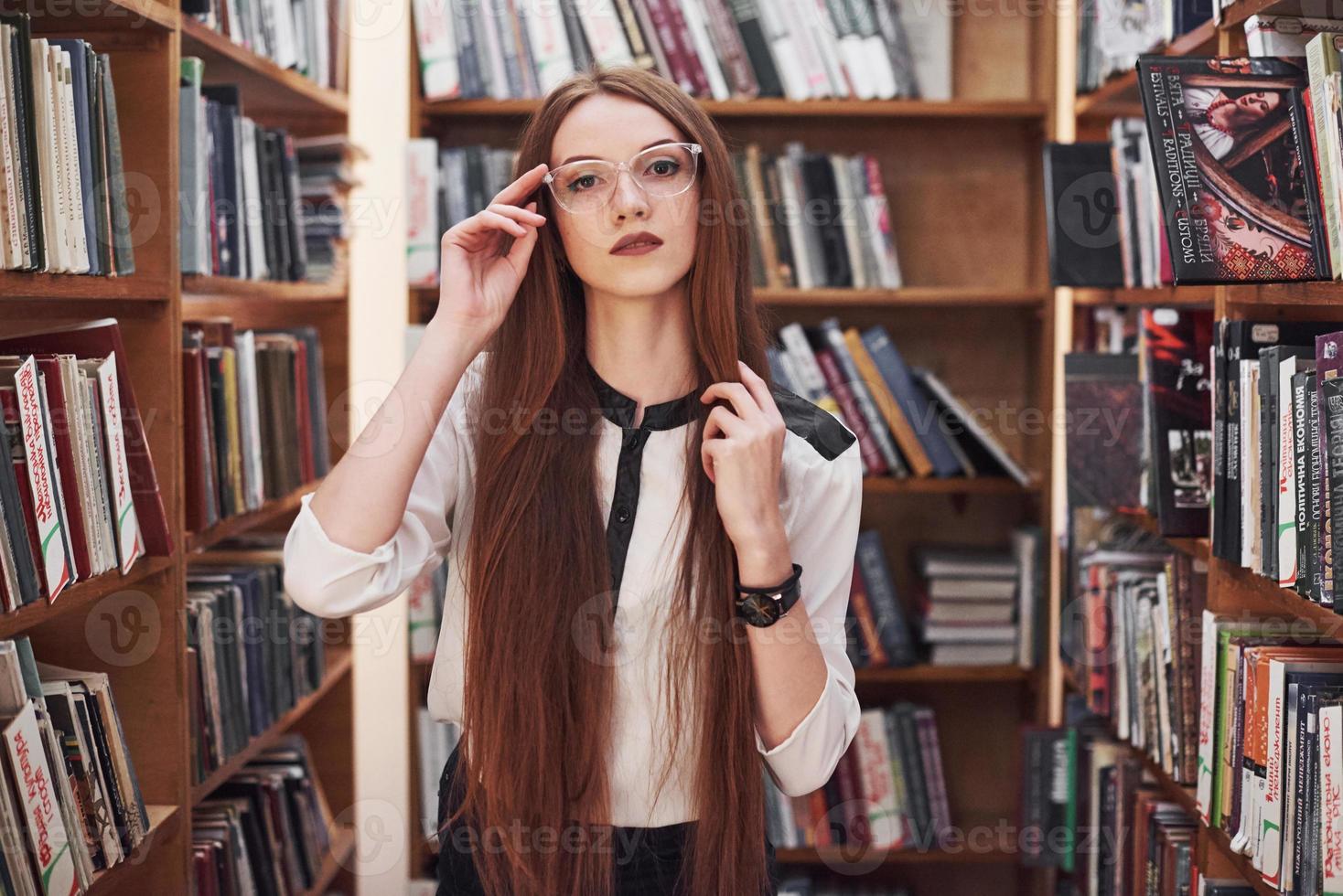 Joven y atractiva bibliotecaria estudiante leyendo un libro entre las estanterías de la biblioteca foto