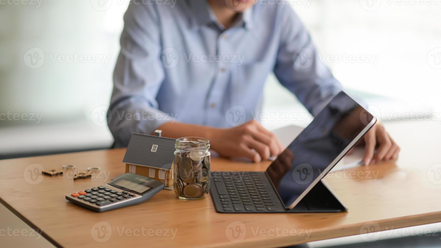 computadora portátil, moneda en una botella de vidrio, calculadora con una casa modelo sobre la mesa y un fondo borroso gente sentada mirando documentos. foto