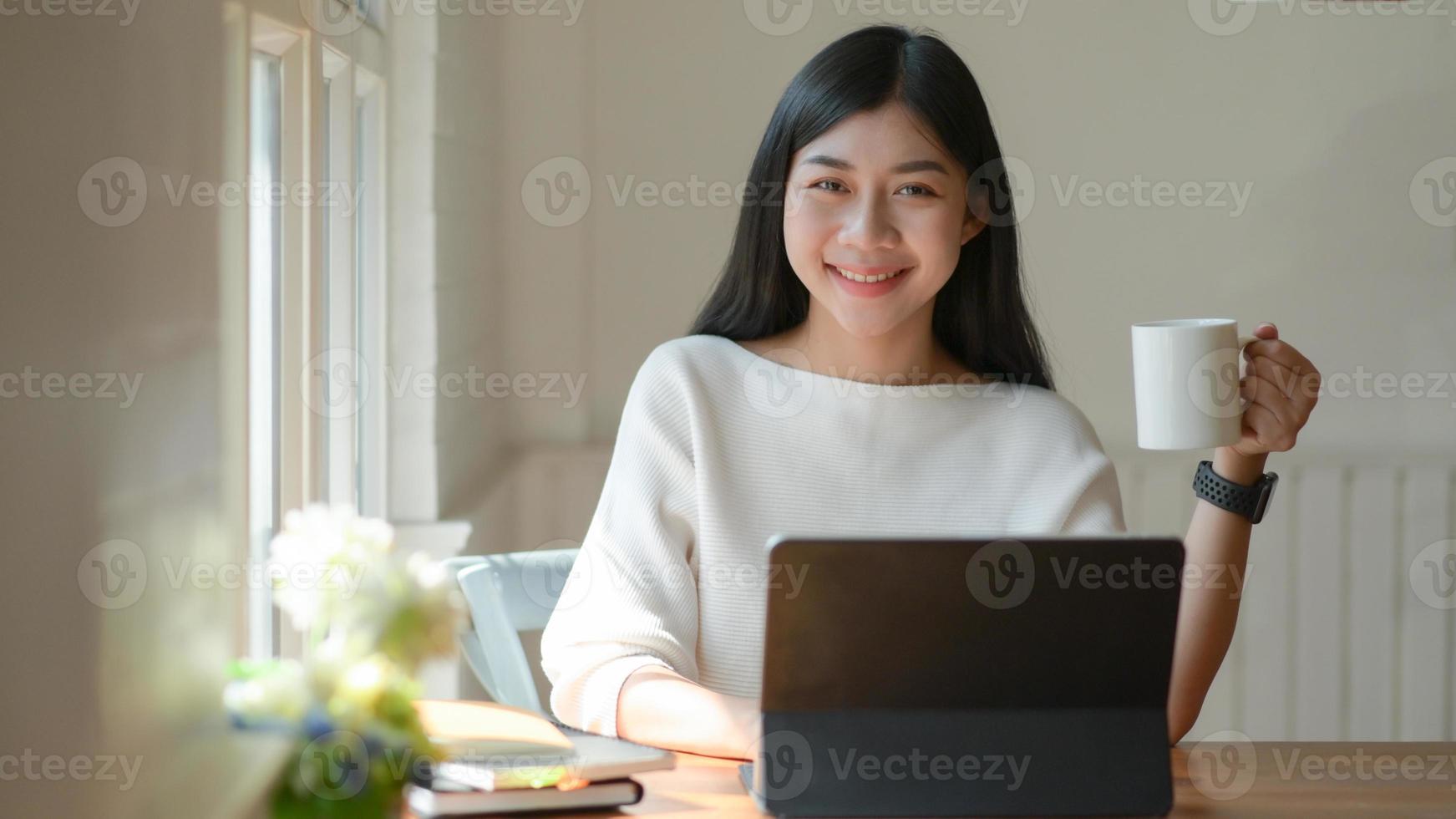 Young girl holds a coffee cup and uses a laptop. She looked at the camera and smiled happily. photo