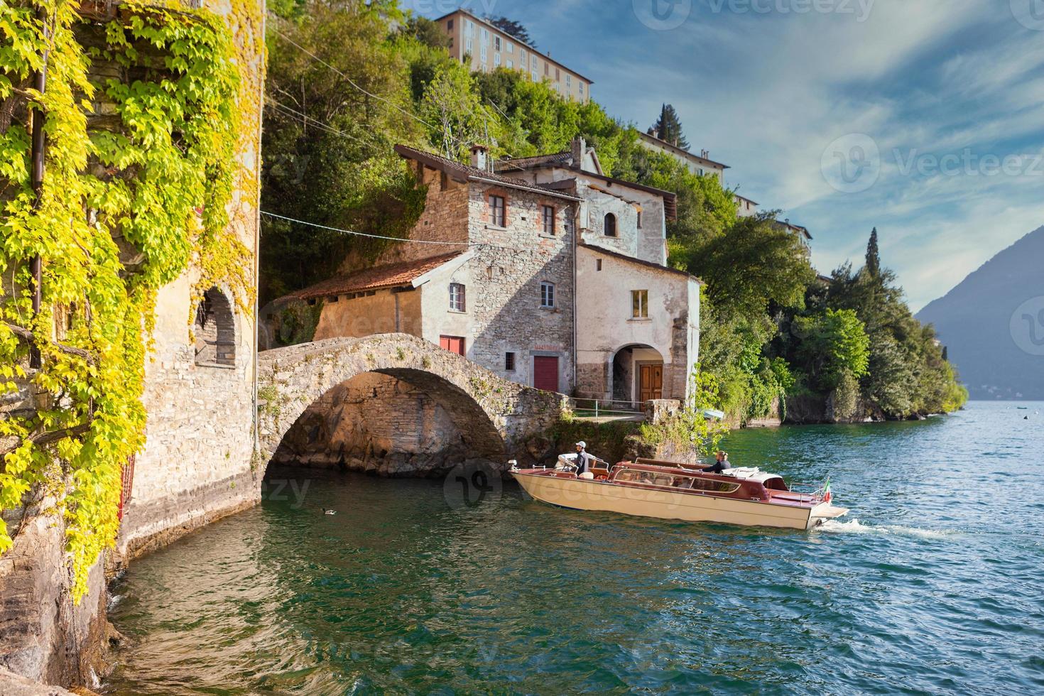 Lancha rápida de madera turística de moda acercándose a un antiguo puente de piedra en el lago de Como foto