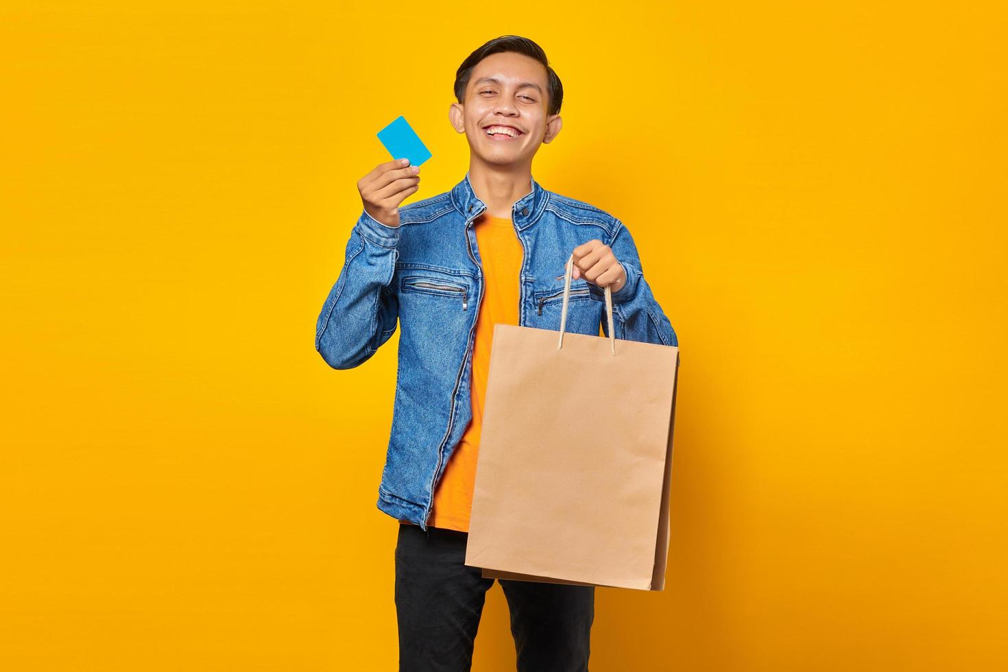 Portrait of cheerful asian man holding shopping bag and showing credit card over yellow background photo