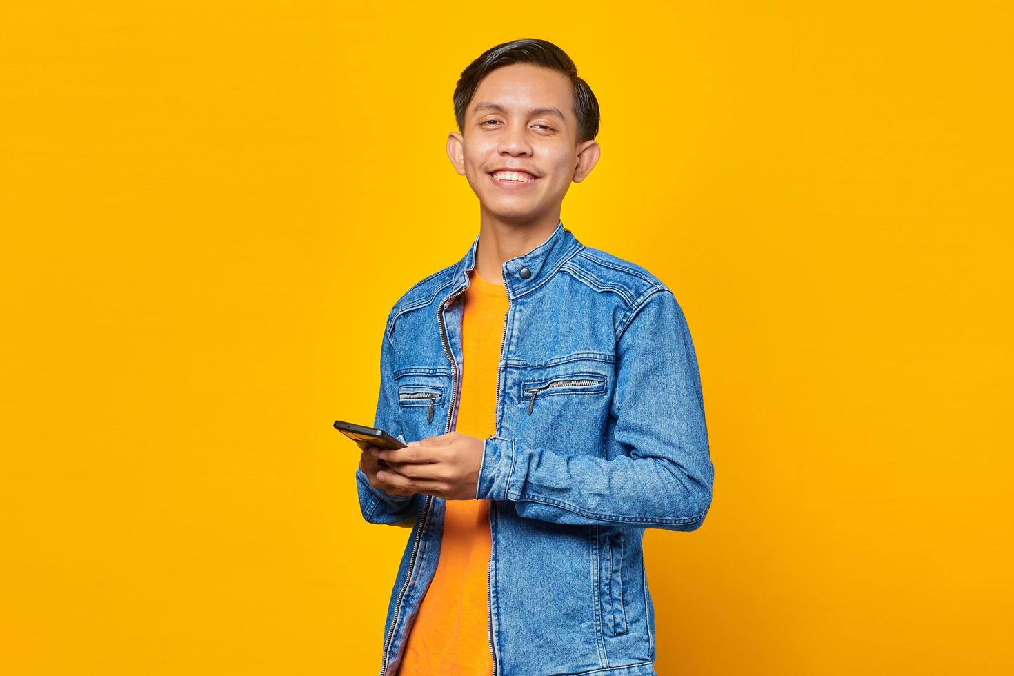 Alegre hombre asiático con smartphone y mirando a la cámara sobre fondo amarillo foto