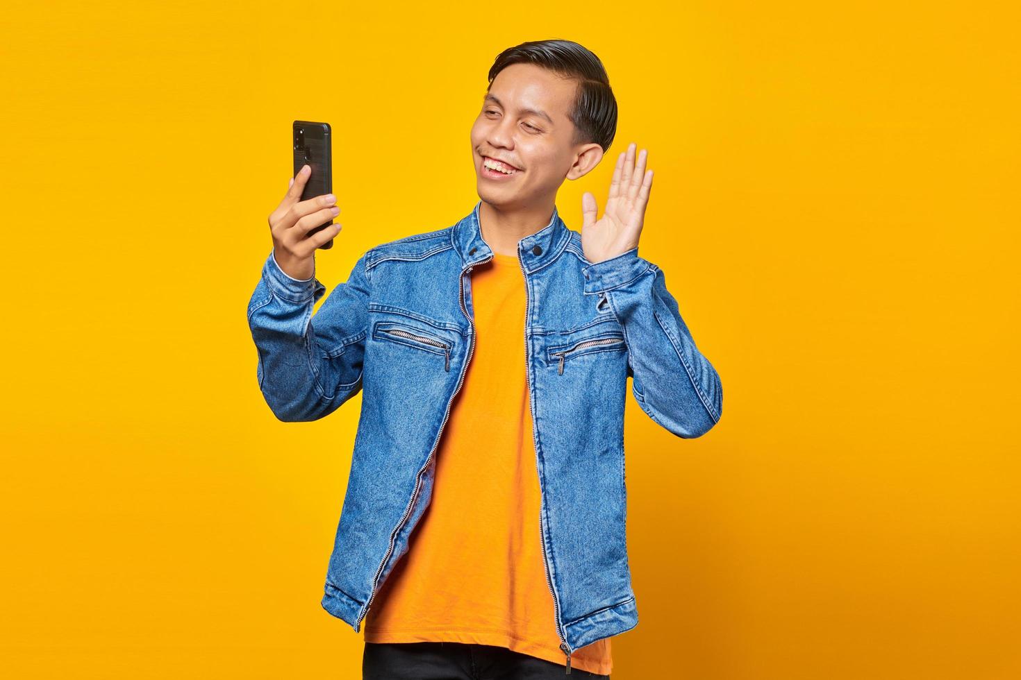 Cheerful asian man looking at smartphone with raised hand over yellow background photo