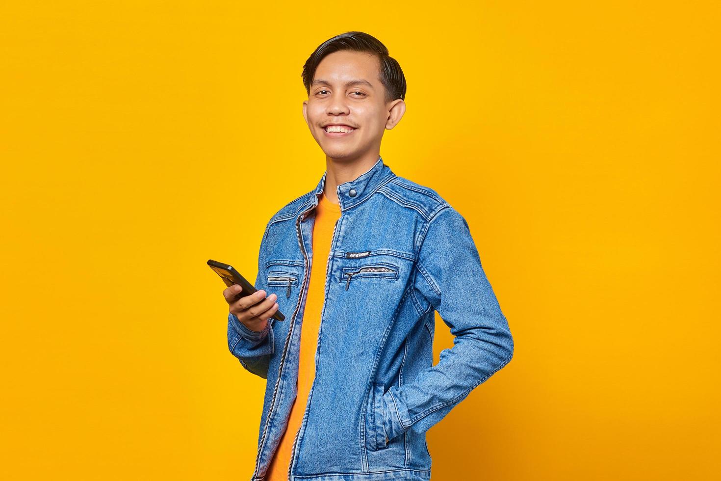 Cheerful asian man using smartphone and looking at camera over yellow background photo