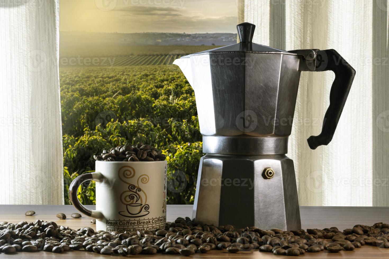 Coffee beans in a cup and Italian coffee maker on a wooden table with coffee field seen through a window in Brazil photo