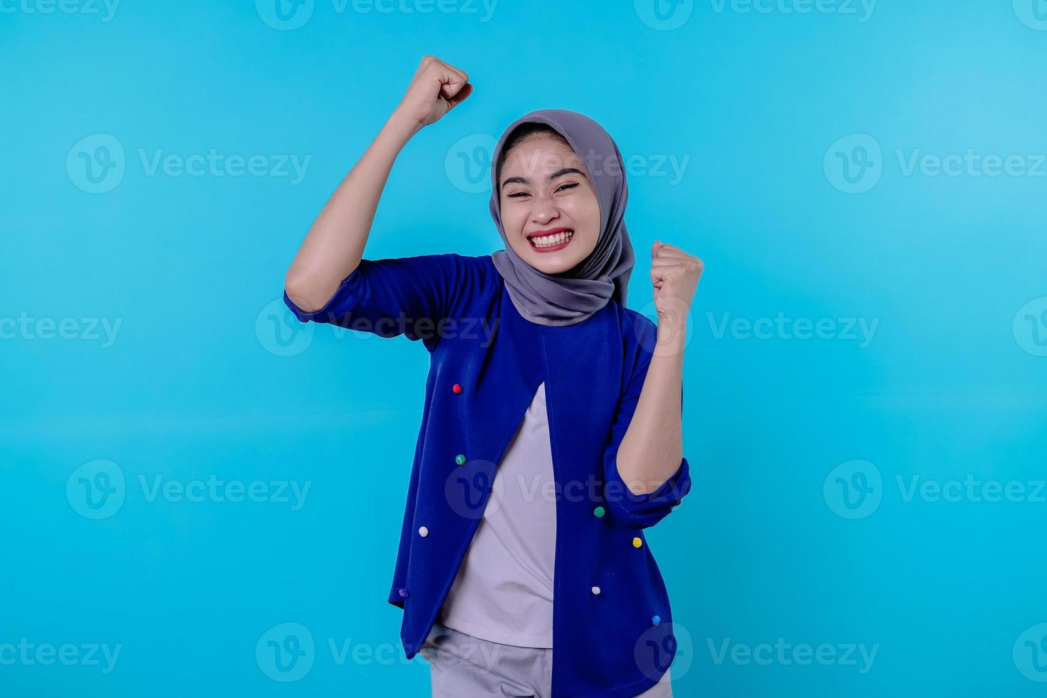 Cheerful young female has positive expression, clenches fists, has overjoyed look, being in high spirit, wearing hijab, isolated over blue wall photo