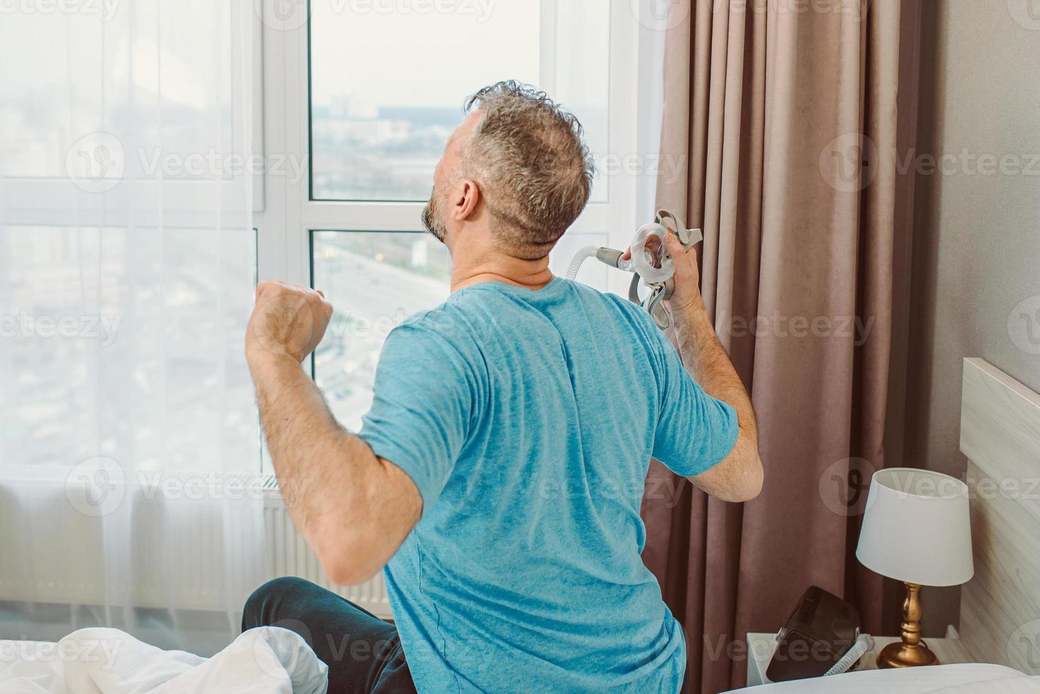 Happy rested man with chronic breathing issues after using  CPAP machine sitting on the bed in bedroom. Healthcare, CPAP, Obstructive sleep apnea therapy, snoring concept photo