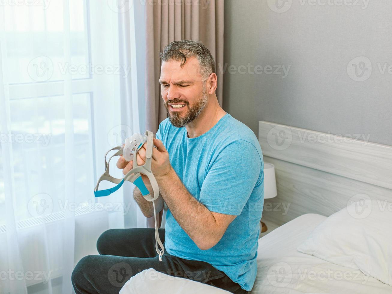Mature happy man with chronic breathing issues considers using CPAP machine sitting on the bed in bedroom . Healthcare, CPAP, snoring concept photo