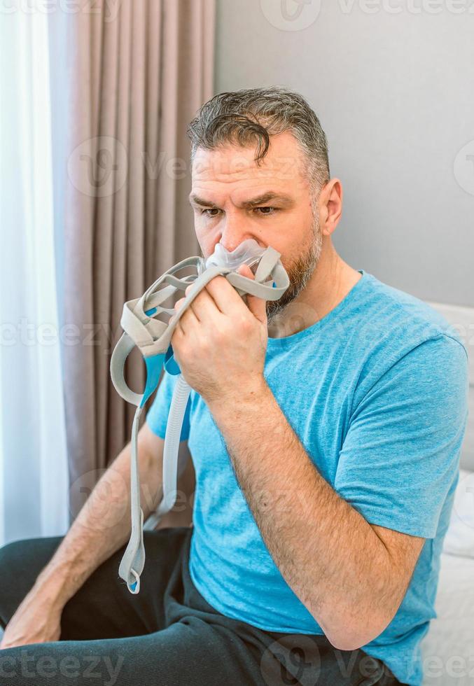 hombre maduro feliz con problemas respiratorios crónicos considera usar sentarse en la cama en el dormitorio con máquina cpap. cuidado de la salud, cpap, concepto de ronquidos foto