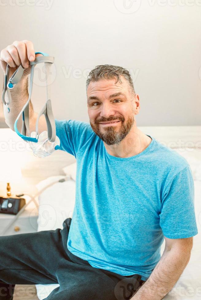 Happy rested man with chronic breathing issues after using  CPAP machine sitting on the bed in bedroom. Healthcare, CPAP, Obstructive sleep apnea therapy, snoring concept photo