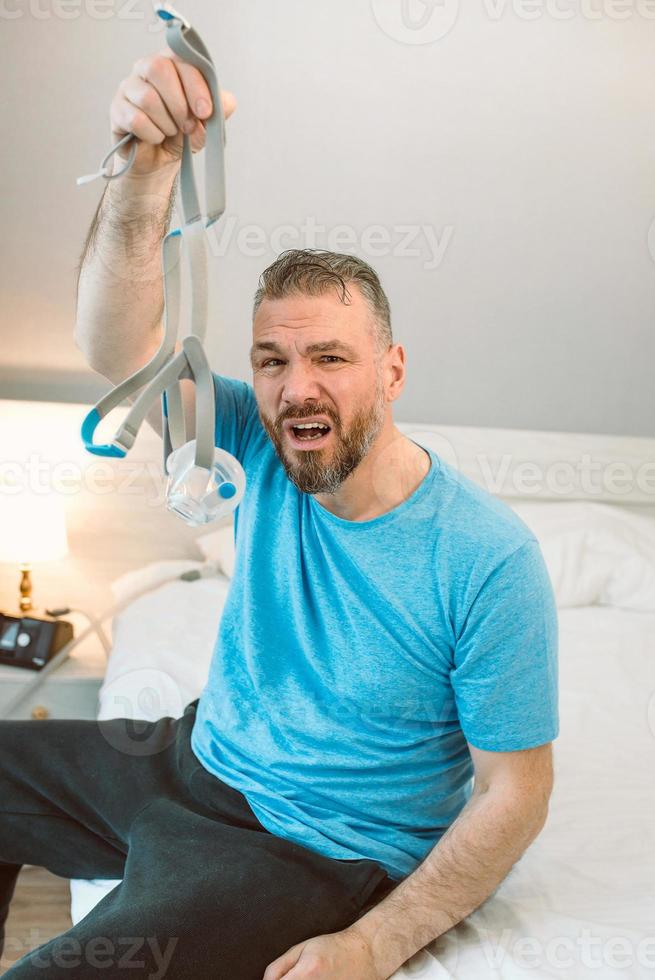 Unhappy shocked man with chronic breathing issues surprised by using  CPAP machine sitting on the bed in bedroom. Healthcare, CPAP, Obstructive sleep apnea therapy, snoring concept photo