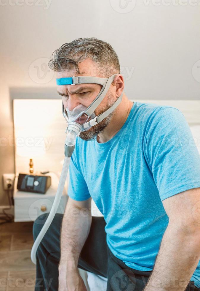 Unhappy shocked man with chronic breathing issues surprised by using  CPAP machine sitting on the bed in bedroom. Healthcare, CPAP, Obstructive sleep apnea therapy, snoring concept photo