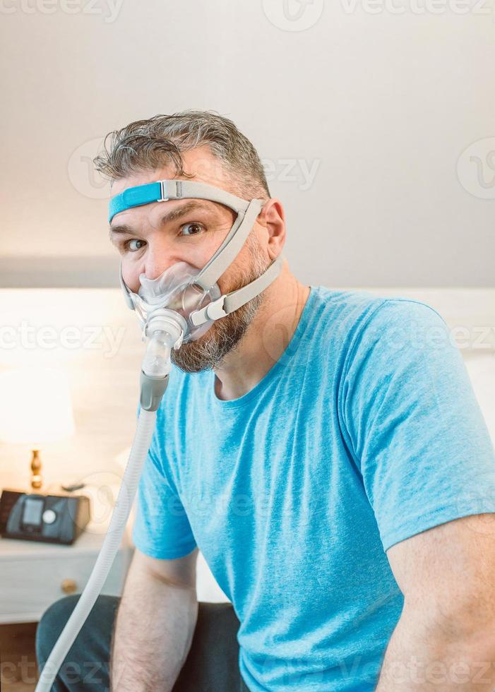 Unhappy shocked man with chronic breathing issues surprised by using  CPAP machine sitting on the bed in bedroom. Healthcare, CPAP, Obstructive sleep apnea therapy, snoring concept photo