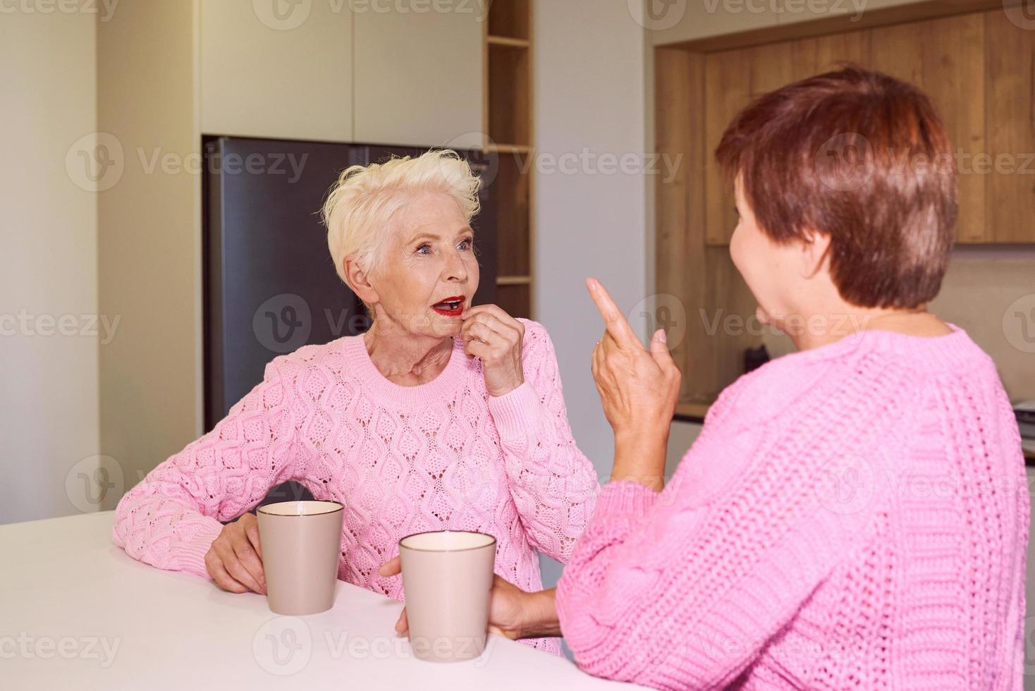 two stylish senior women in pink sweaters sitting with mugs at modern kitchen gossiping. Friendship, talk, gossip, relationships, news, family concept photo